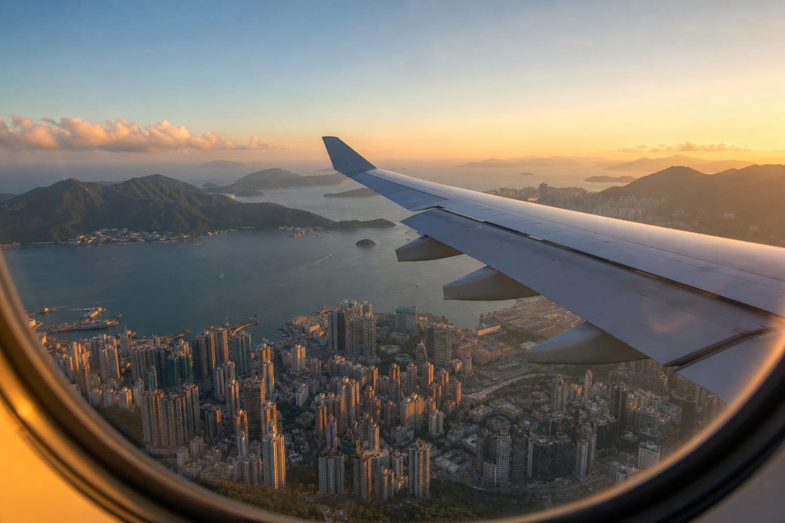 Airplane wing climbing over a harbor city with green mountains at golden hour